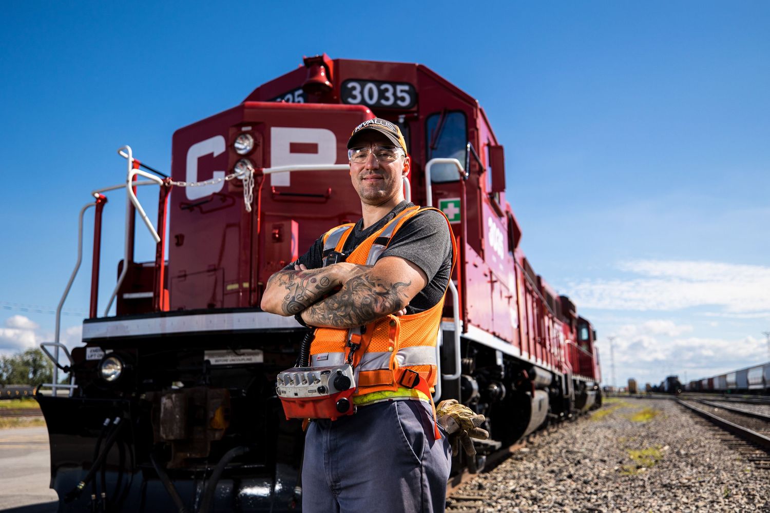 Portrait d'un fier employé du Canadien Pacifique devant des trains rouges alignés sur les voies ferrées sous un ciel bleu, à Montréal.