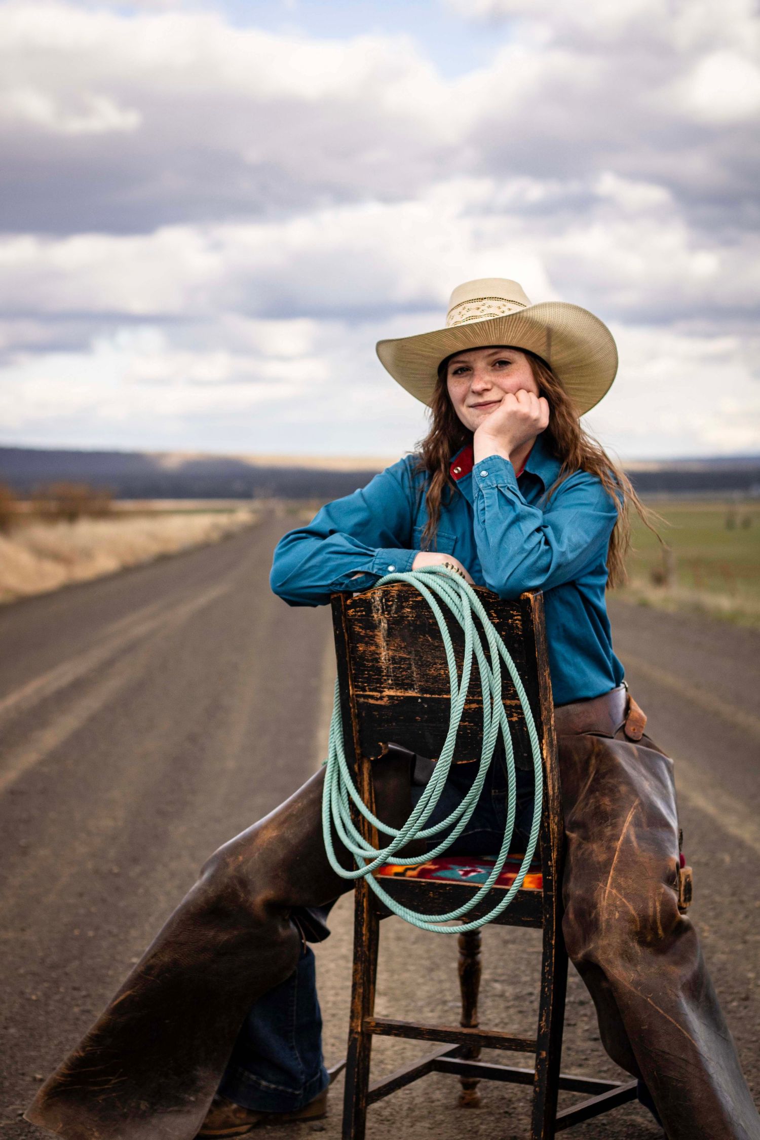 young woman sits on backwards chair with rope, chaps, and cowboy hat. behind her is a long dirt road, hills, and a cloudy sky