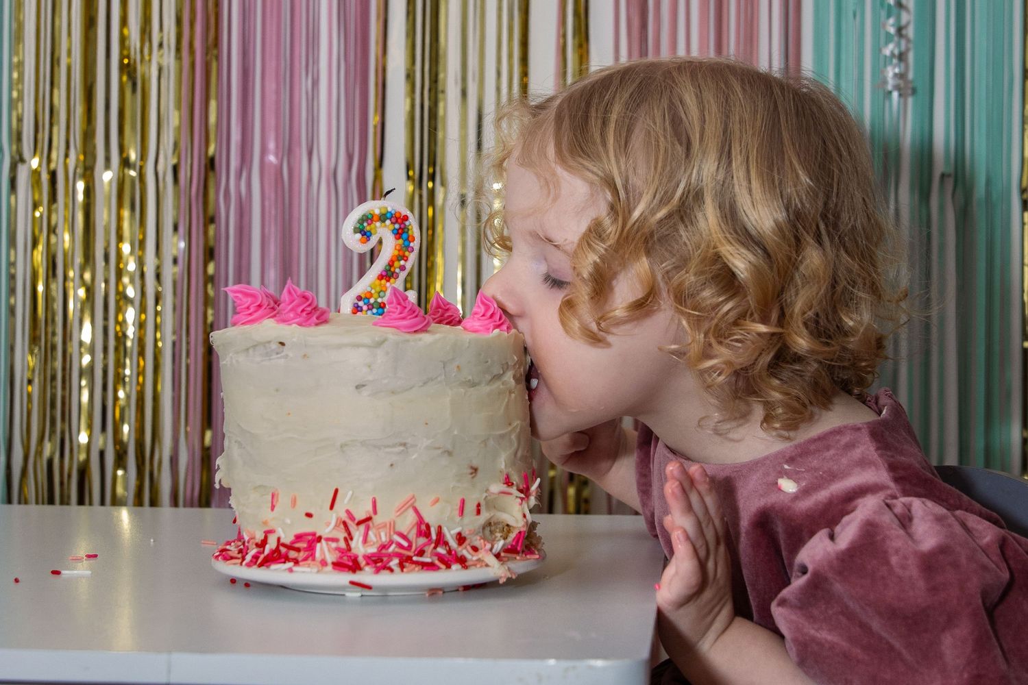 Child with curly hair blowing out a birthday candle on a white frosted cake with pink decorations and number 2 topper.
