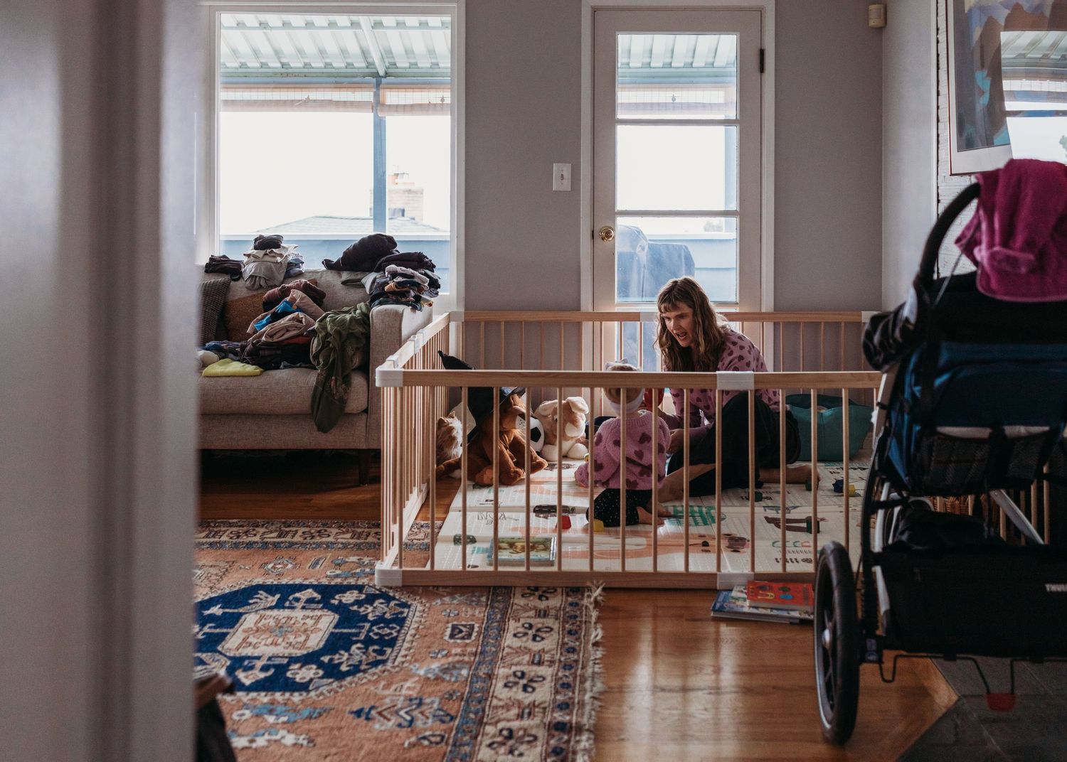 A cozy living room space with a child sitting inside a white baby gate near a patterned carpet and windows.