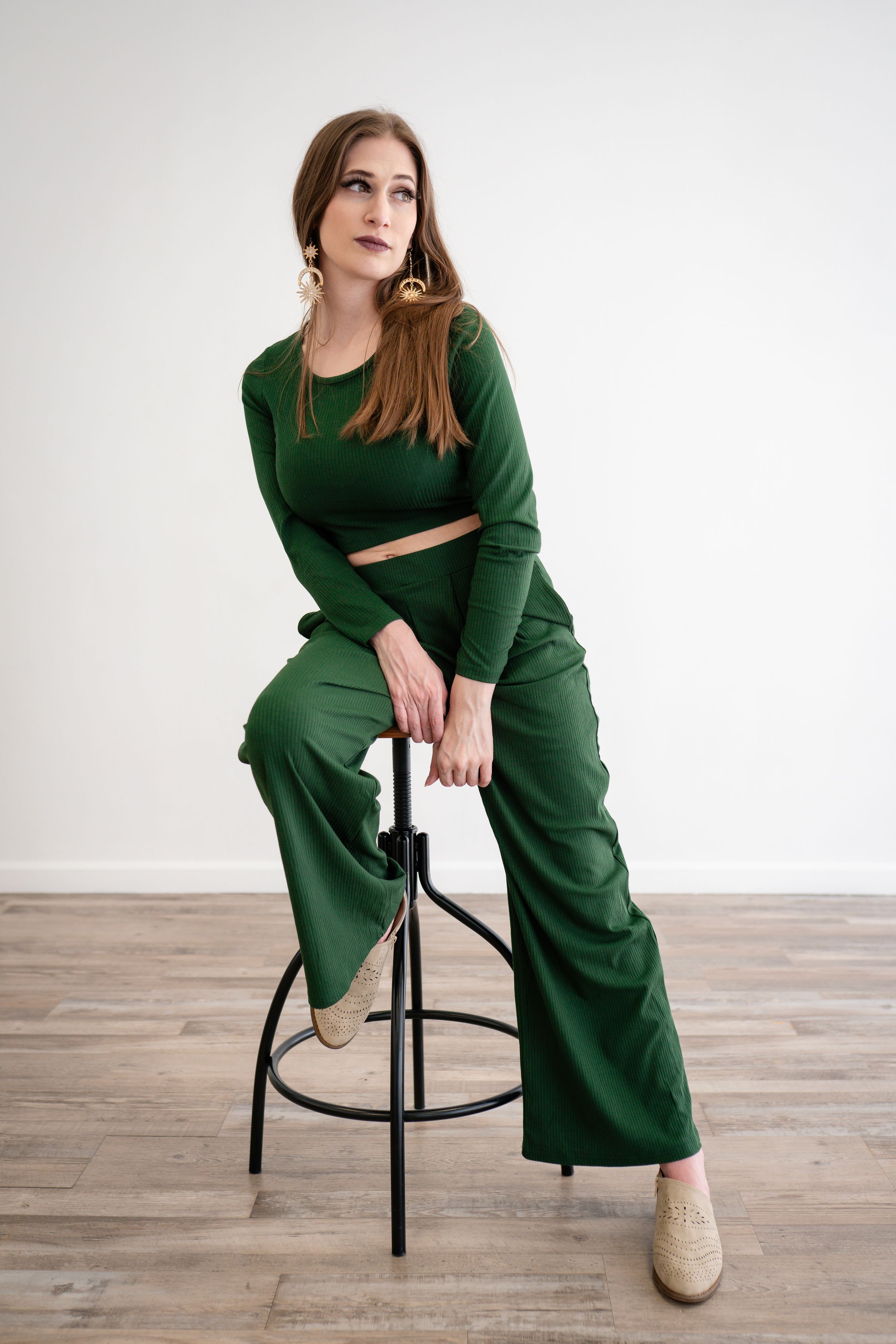 Model wearing emerald green two-piece outfit sits elegantly on black bar stool against white background.