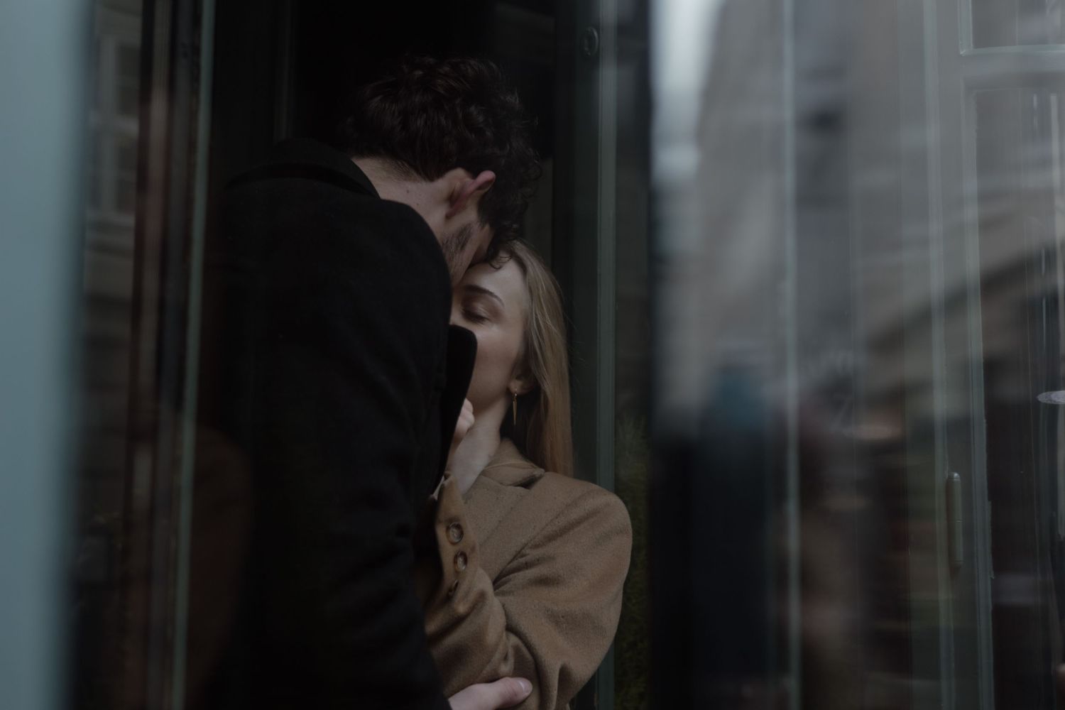 A person wearing a beige coat leans against a window in a dark moody setting with city reflections.