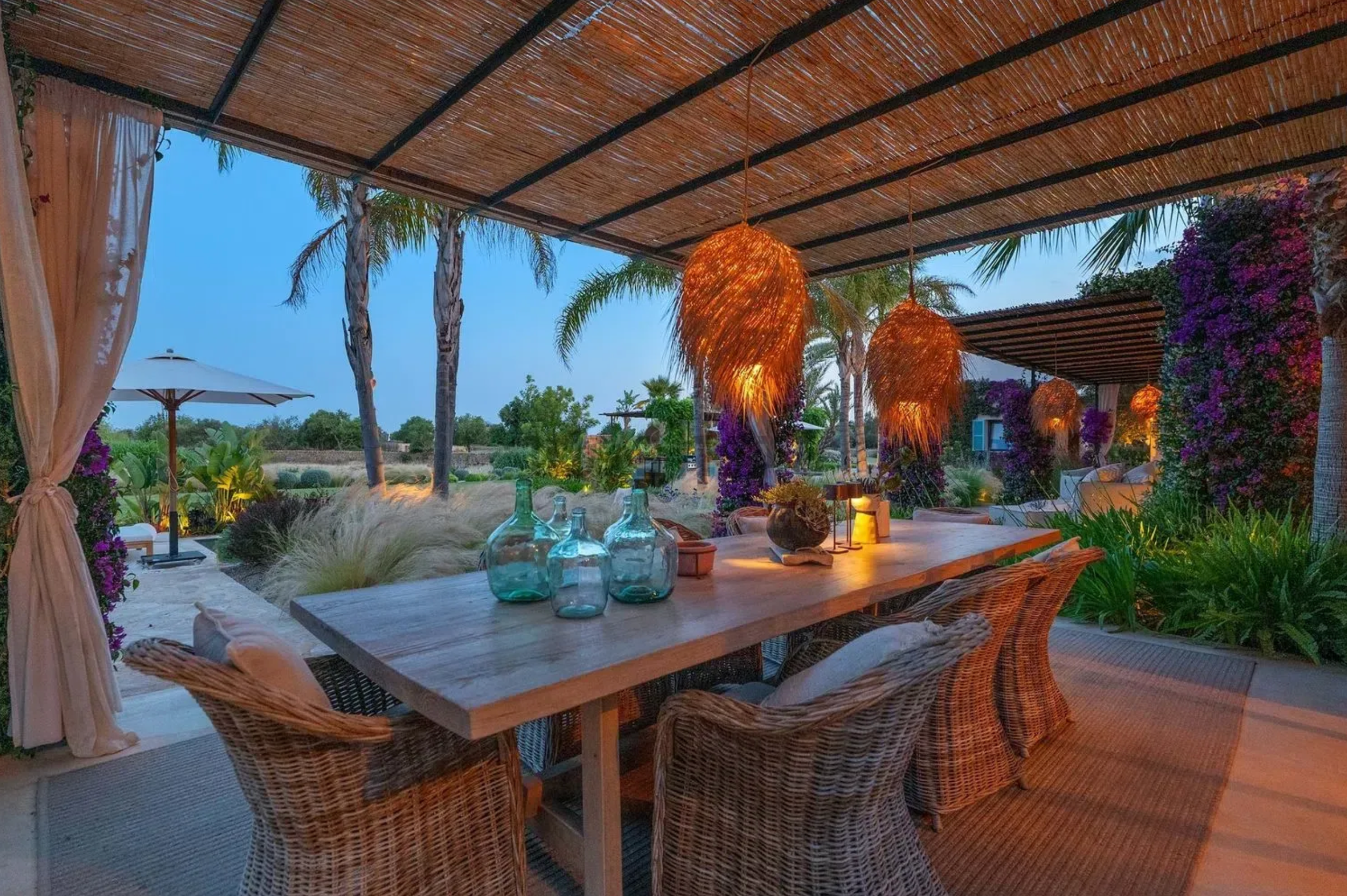 Outdoor dining terrace with wicker chairs and orange lanterns overlooking tropical garden and ocean view at sunset.