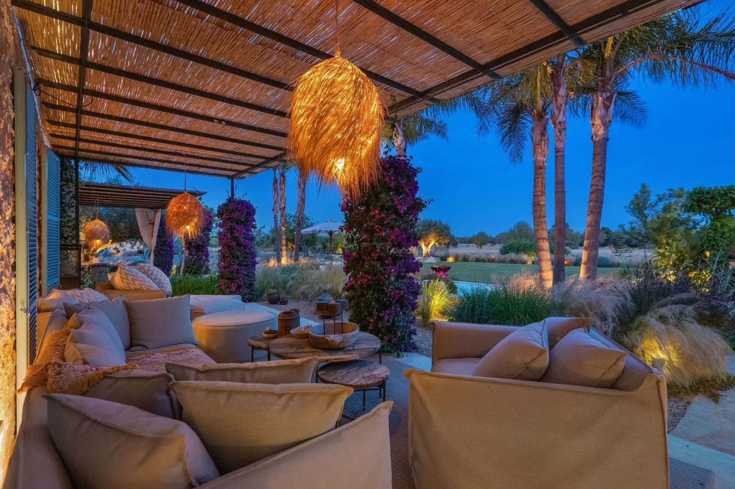 Outdoor covered patio with comfortable lounge seating and warm lighting overlooking a tropical garden.