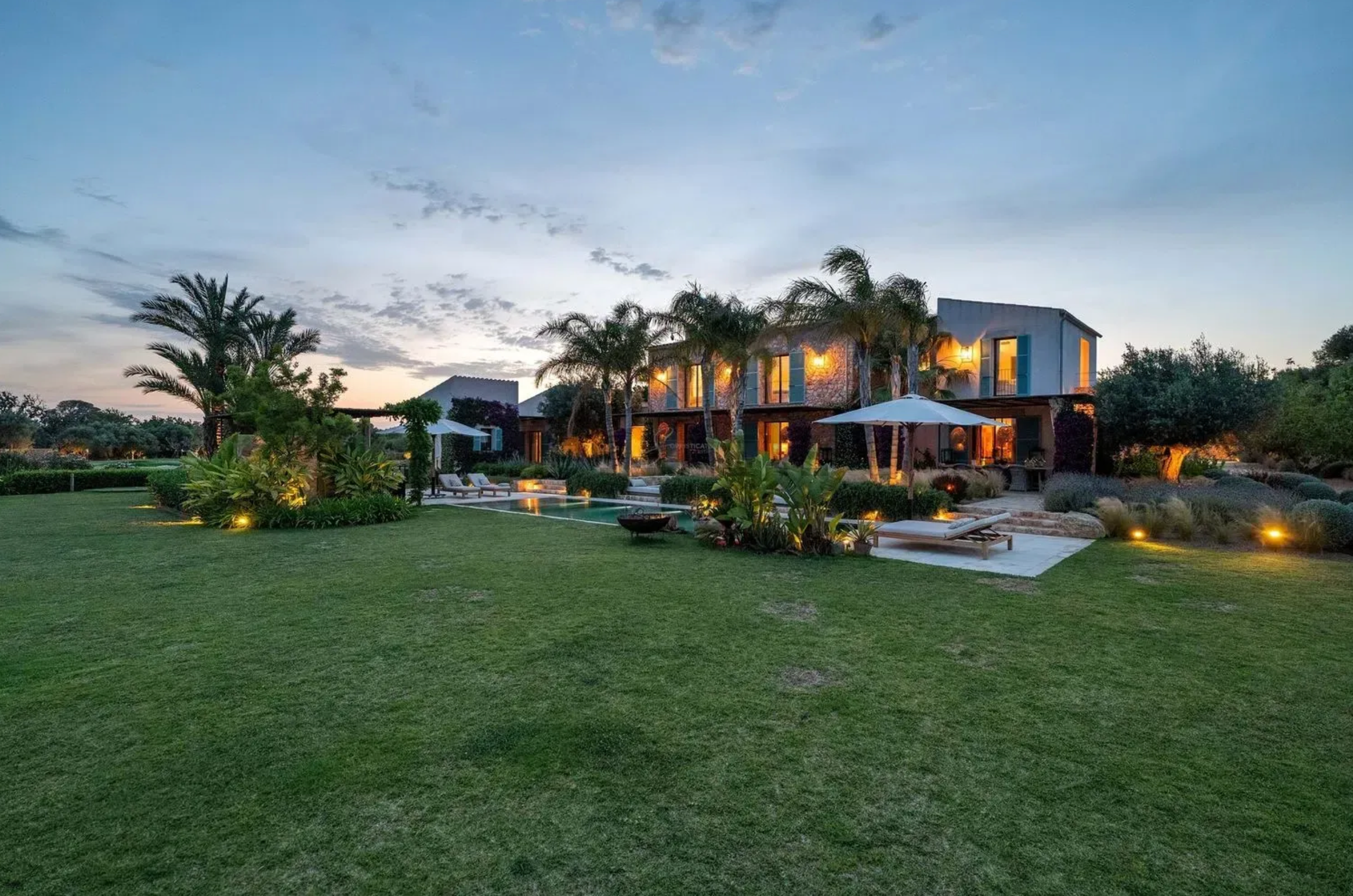 Modern luxury villa with infinity pool illuminated at dusk against a dramatic sunset sky.