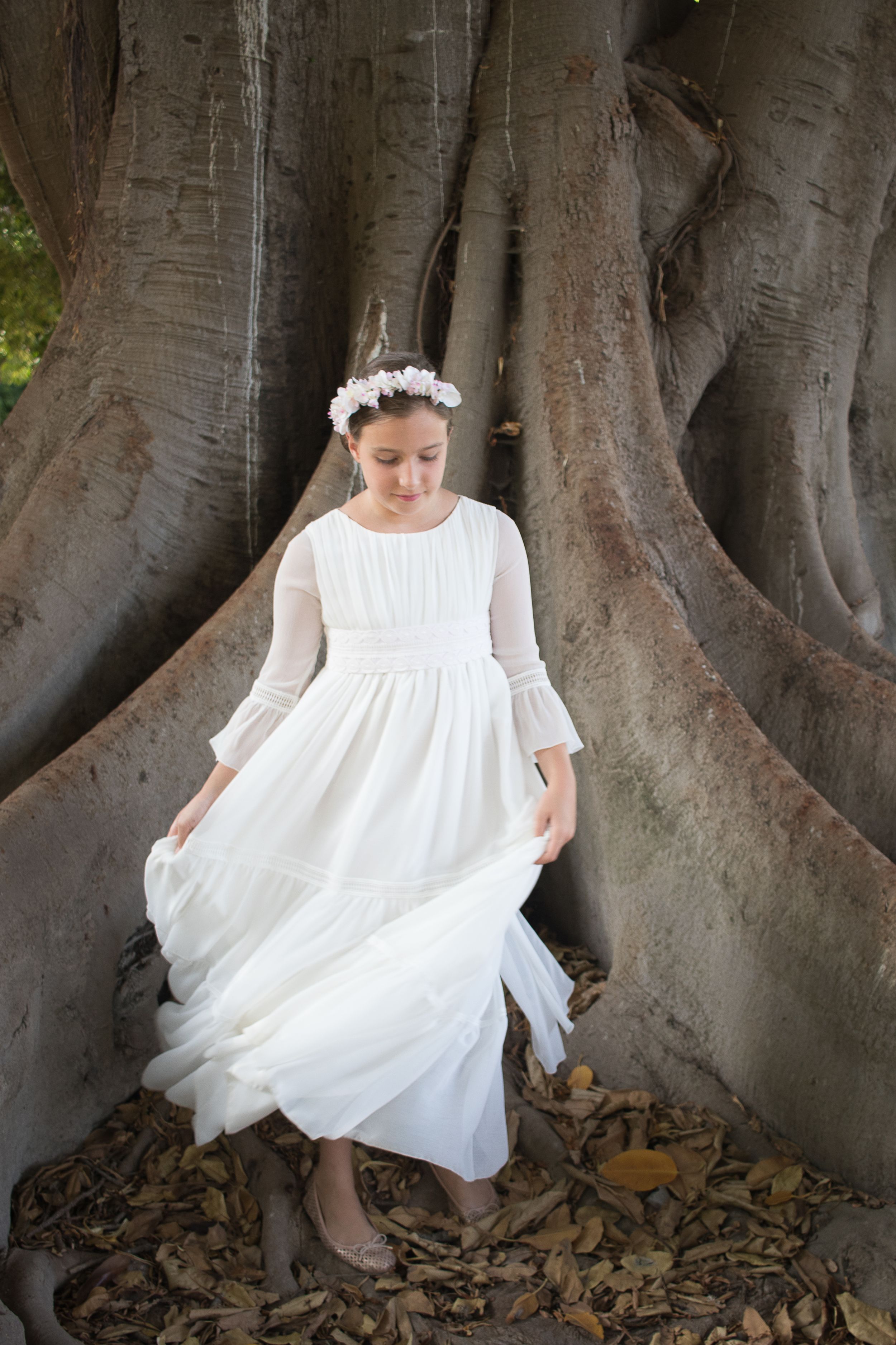 A young figure in a flowing white dress and floral crown poses against the massive roots of an ancient tree.