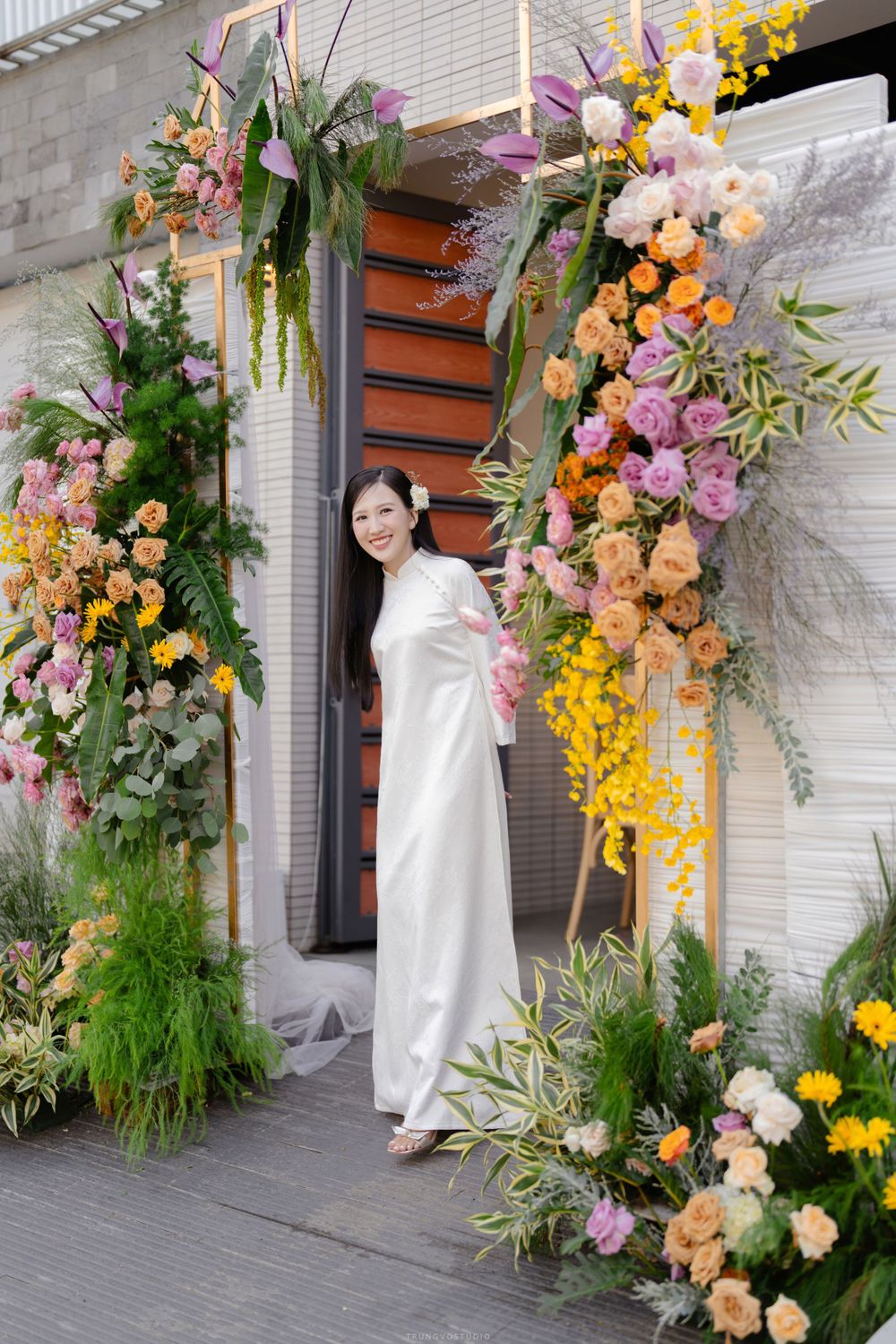 Bride in white ao dai smiles under colorful floral arch at wedding ceremony entrance.