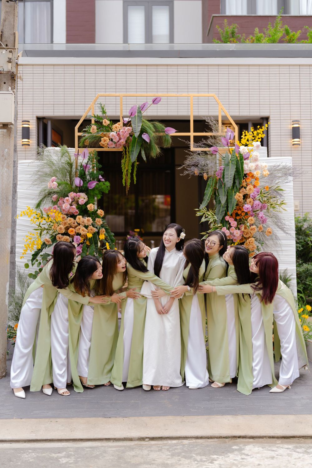 Bride surrounded by smiling bridesmaids in green ao dai under colorful floral arch.