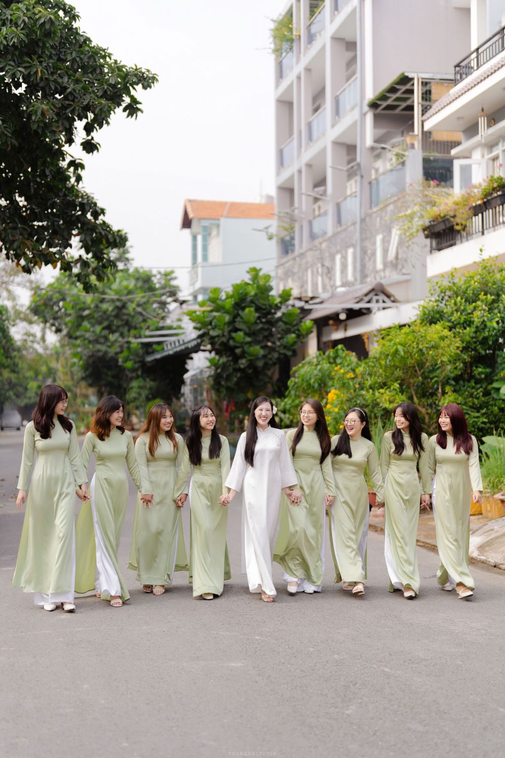 Bride in white ao dai walks with bridesmaids in matching sage green dresses on street.