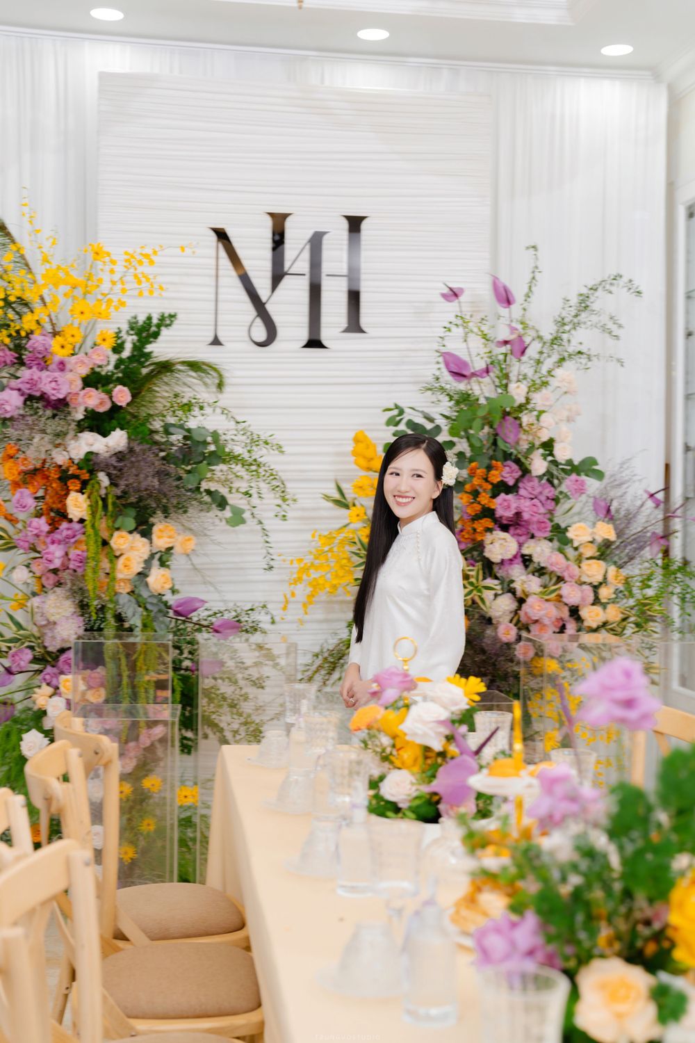 Smiling bride in white ao dai stands among colorful floral arrangements at decorated venue.
