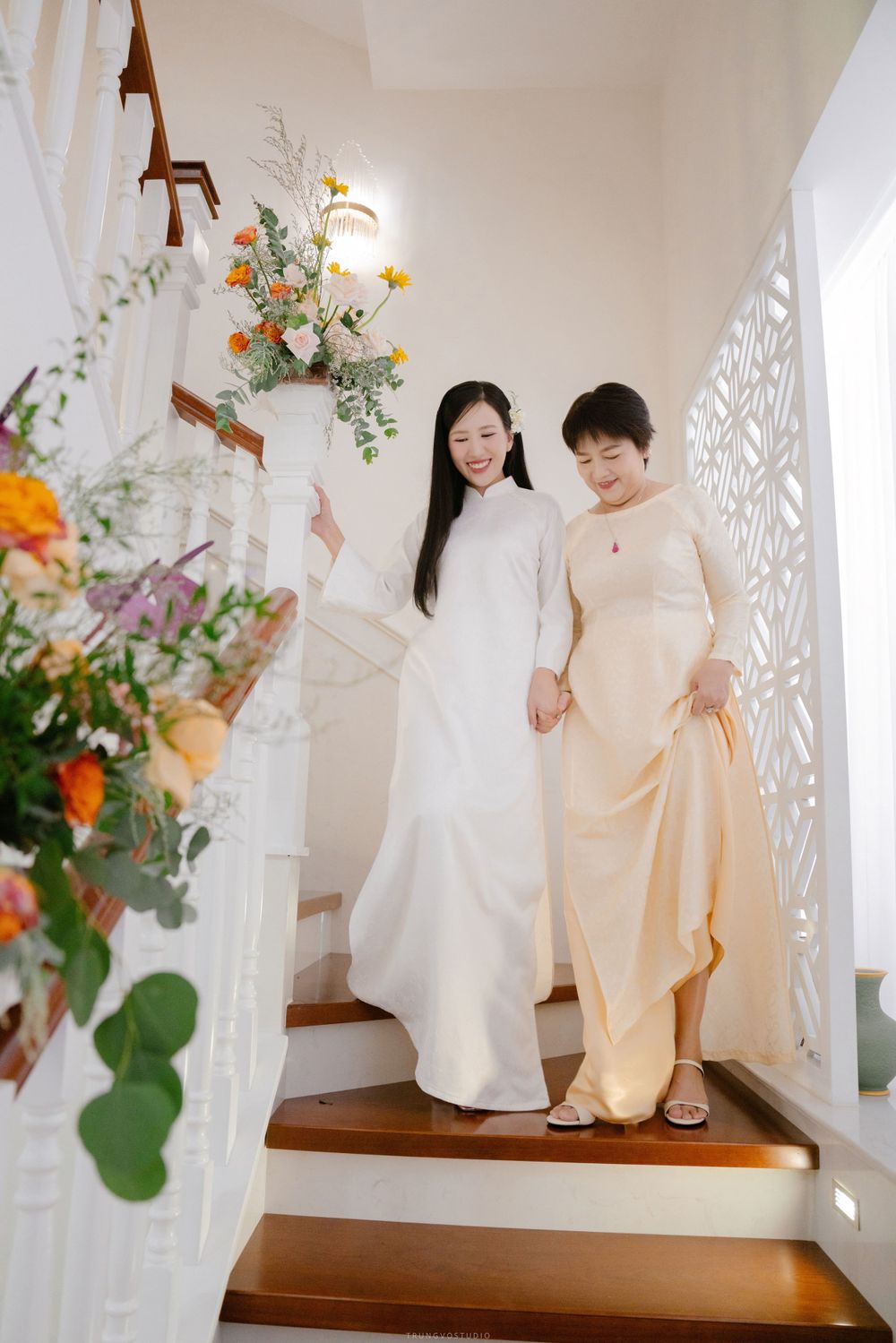 Bride in white ao dai descends staircase with mother at traditional Vietnamese wedding.
