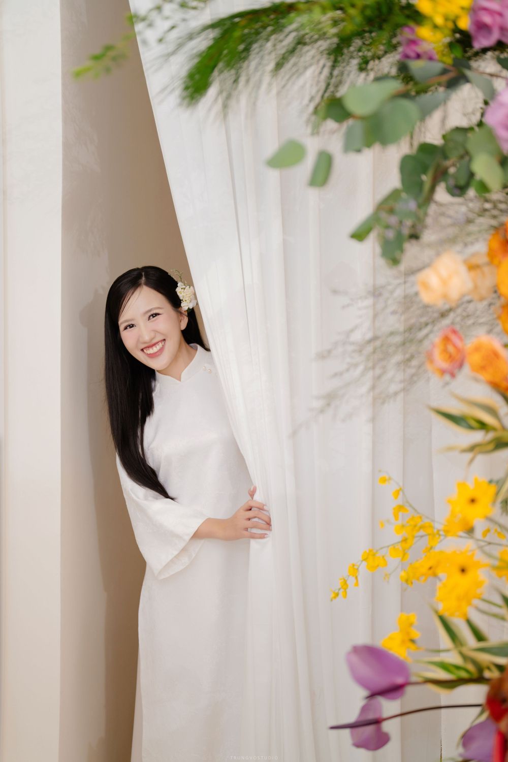 Bride in white ao dai peeks from behind curtain surrounded by colorful floral decorations.