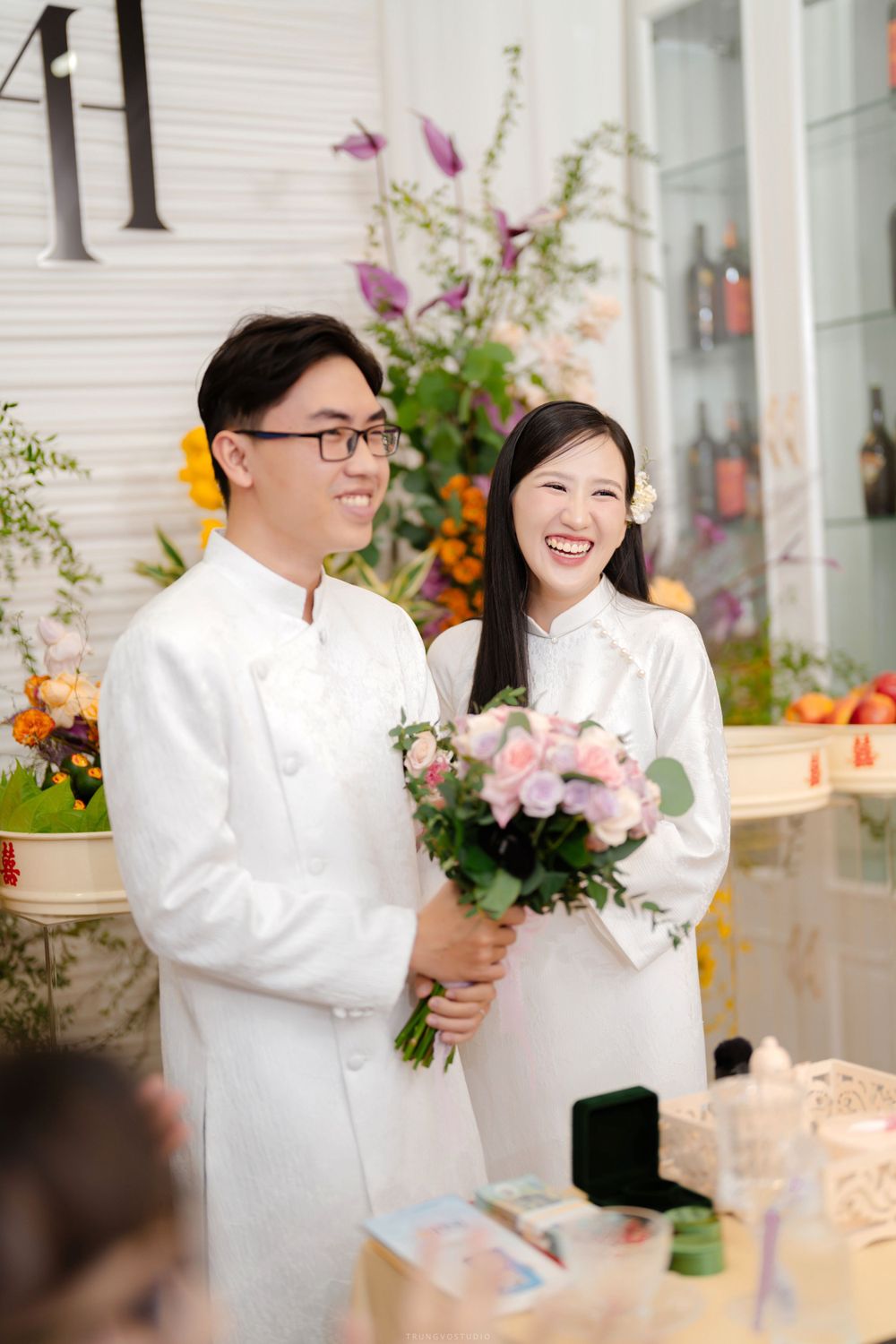 Smiling couple in matching white ao dai hold pink bouquet at traditional Vietnamese wedding.