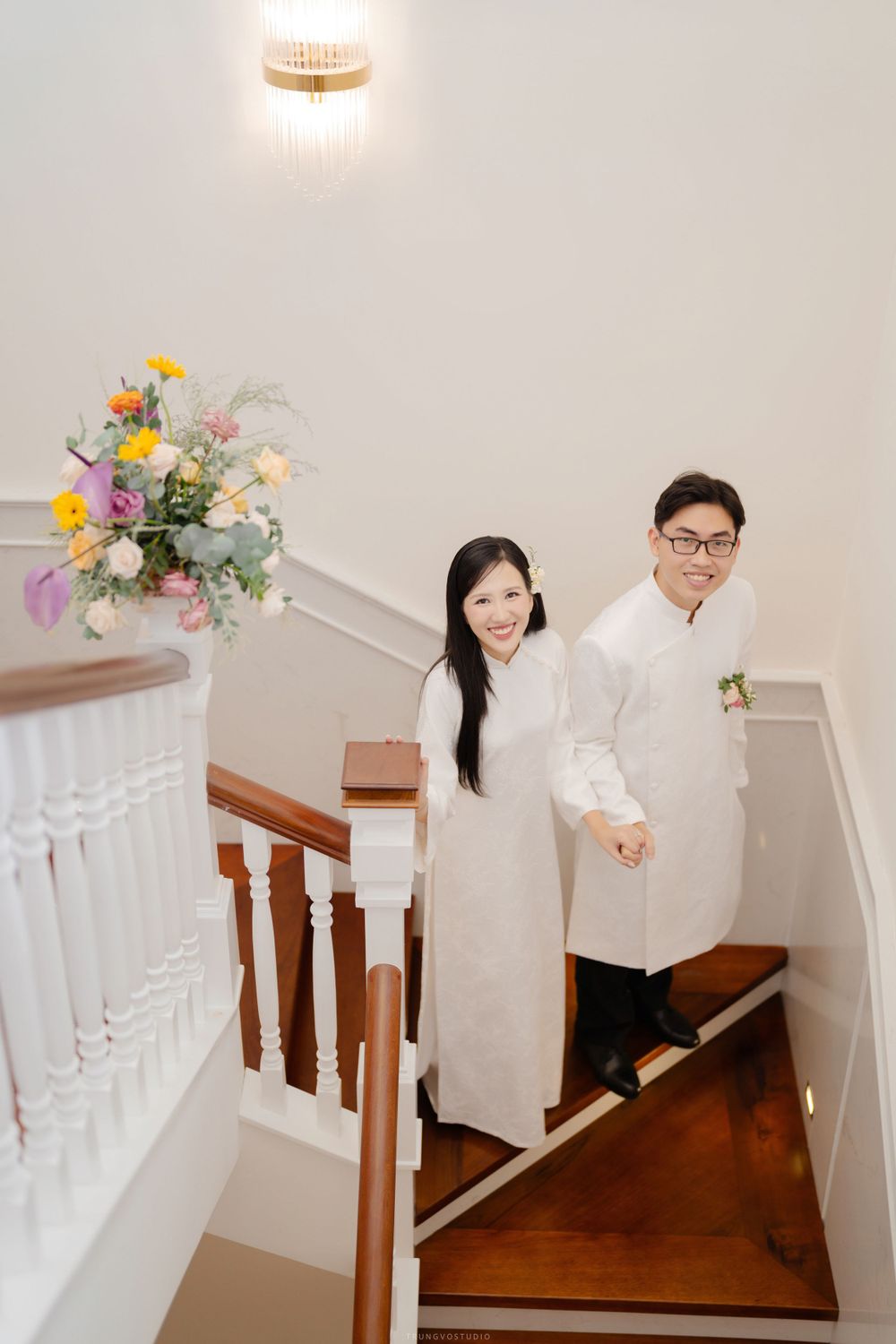 Couple in white ao dai traditional Vietnamese attire smiling on wooden staircase with colorful floral bouquet.