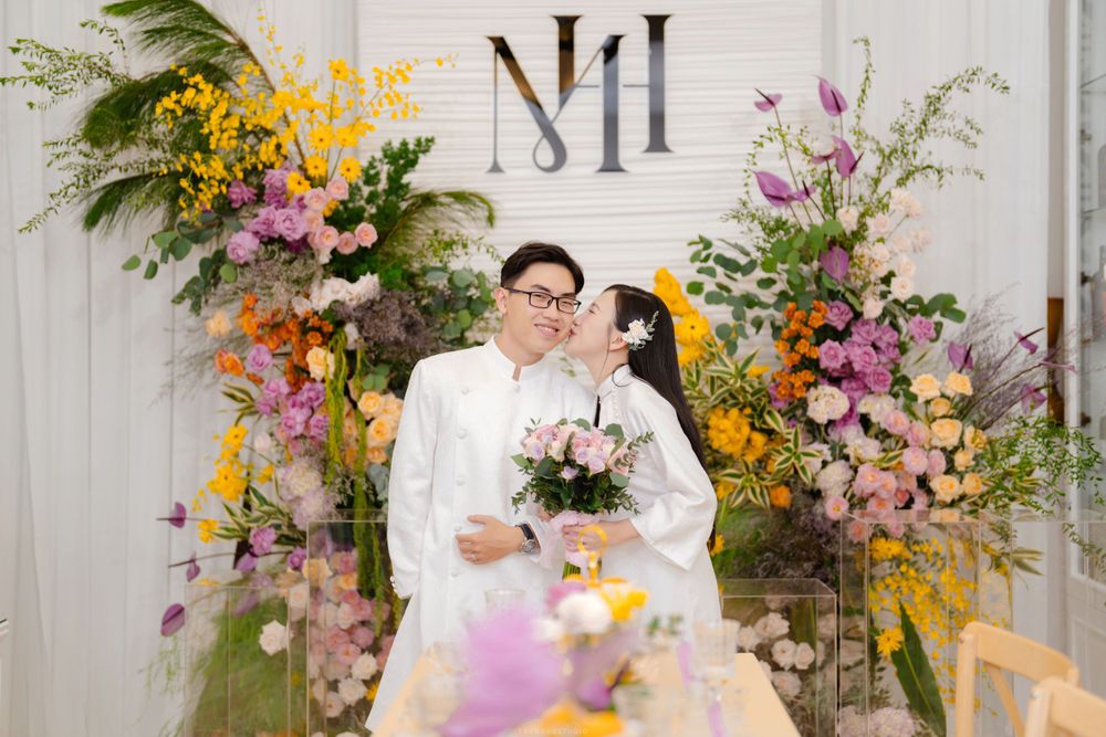 Vietnamese couple in white ao dai kissing at wedding ceremony with colorful floral backdrop and monogram.