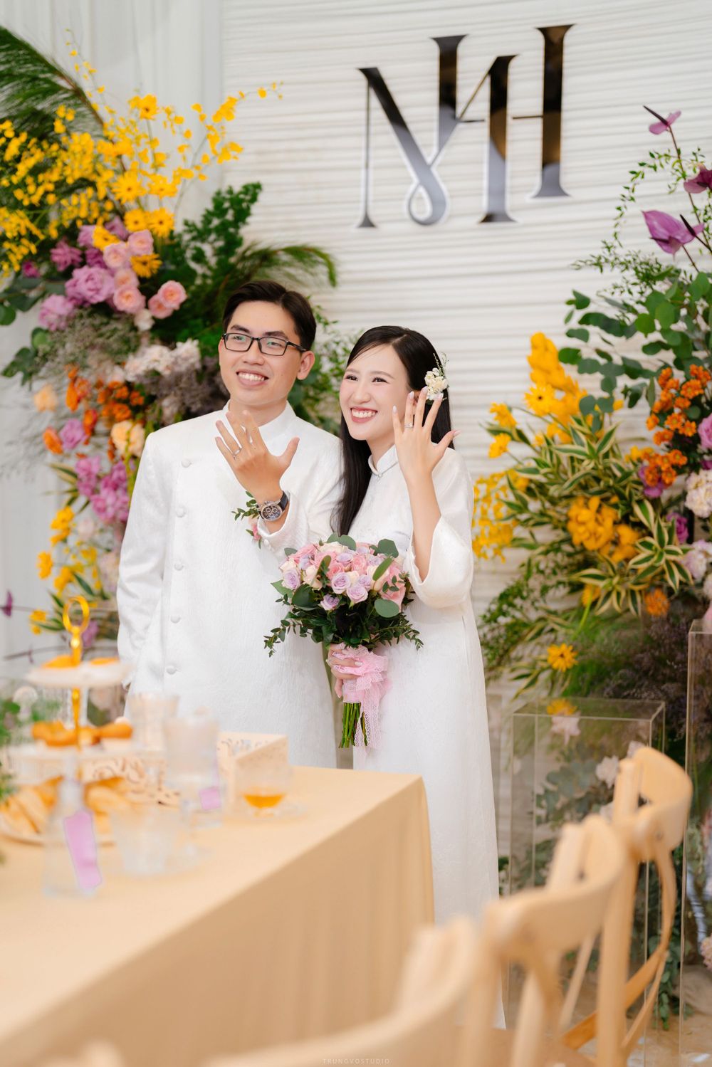 Vietnamese couple in white ao dai showing wedding rings while smiling at decorated ceremony venue.