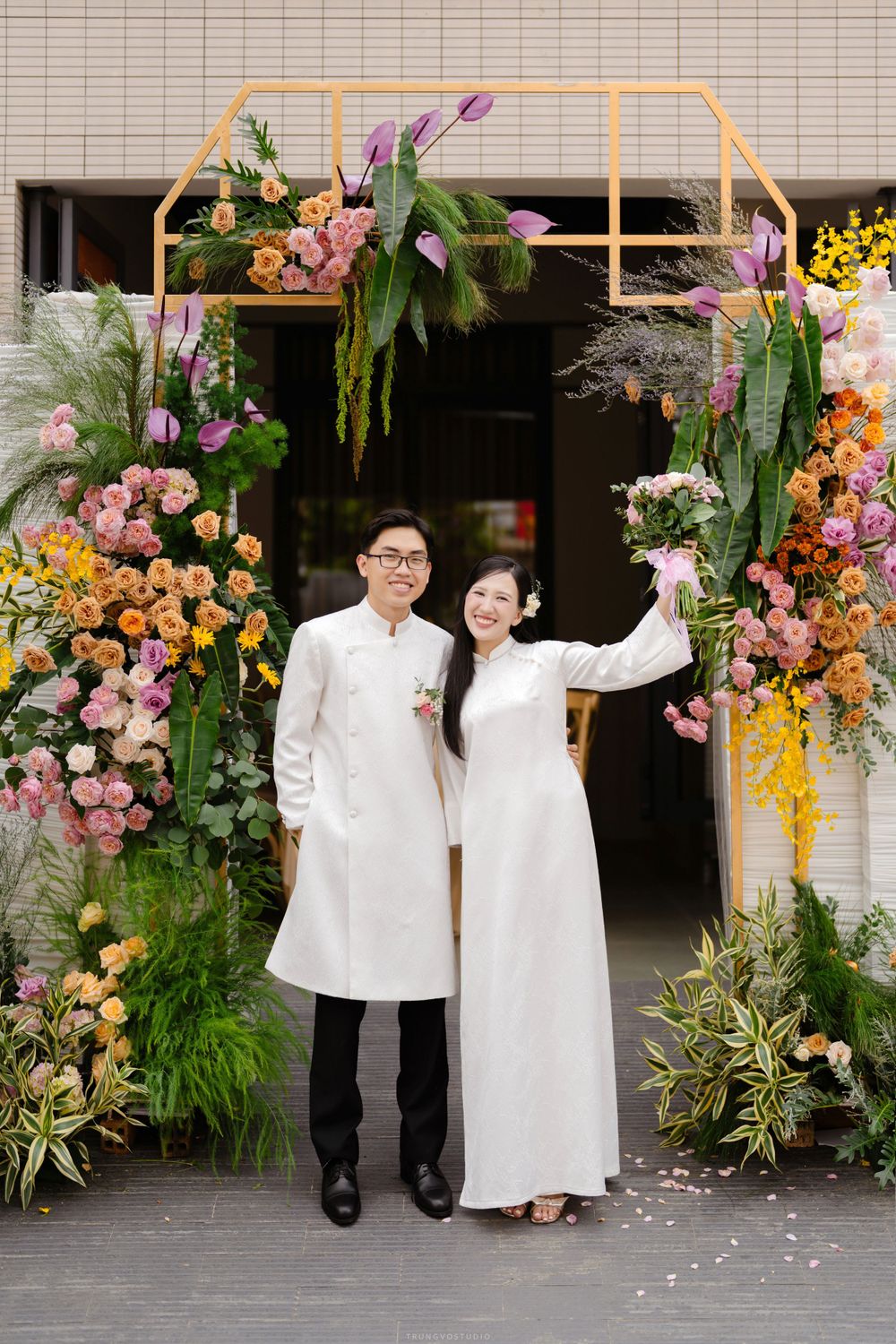 Vietnamese couple in white ao dai posing under colorful floral arch with tropical flowers at wedding.