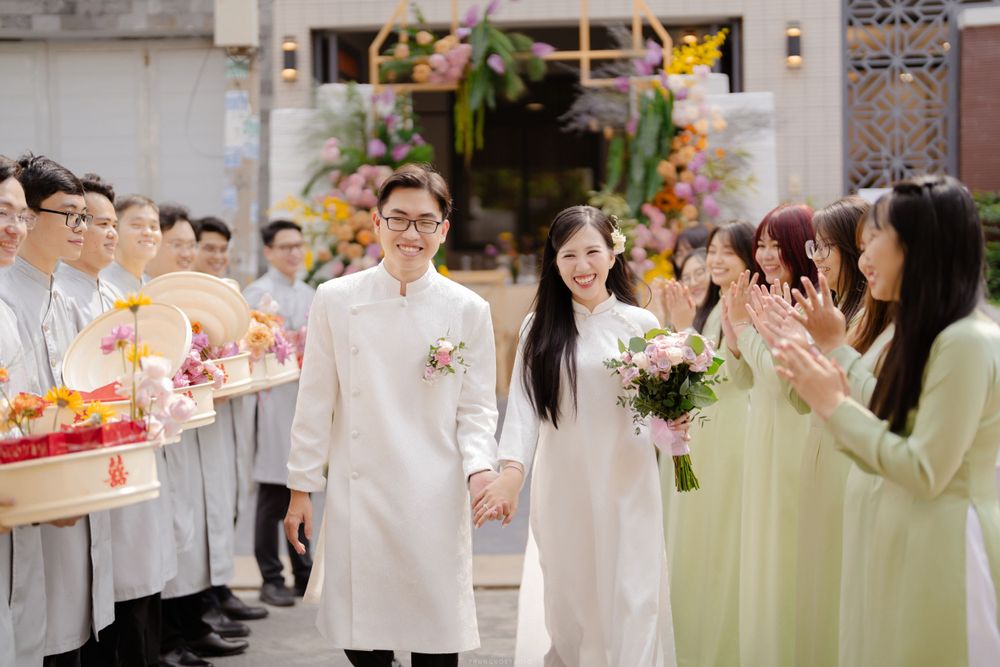 Vietnamese wedding couple in white ao dai walking hand in hand through applauding guests at ceremony.