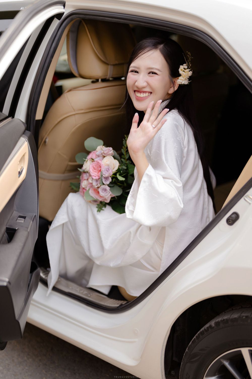 Smiling bride in white ao dai waving from car window while holding pink and white bouquet.