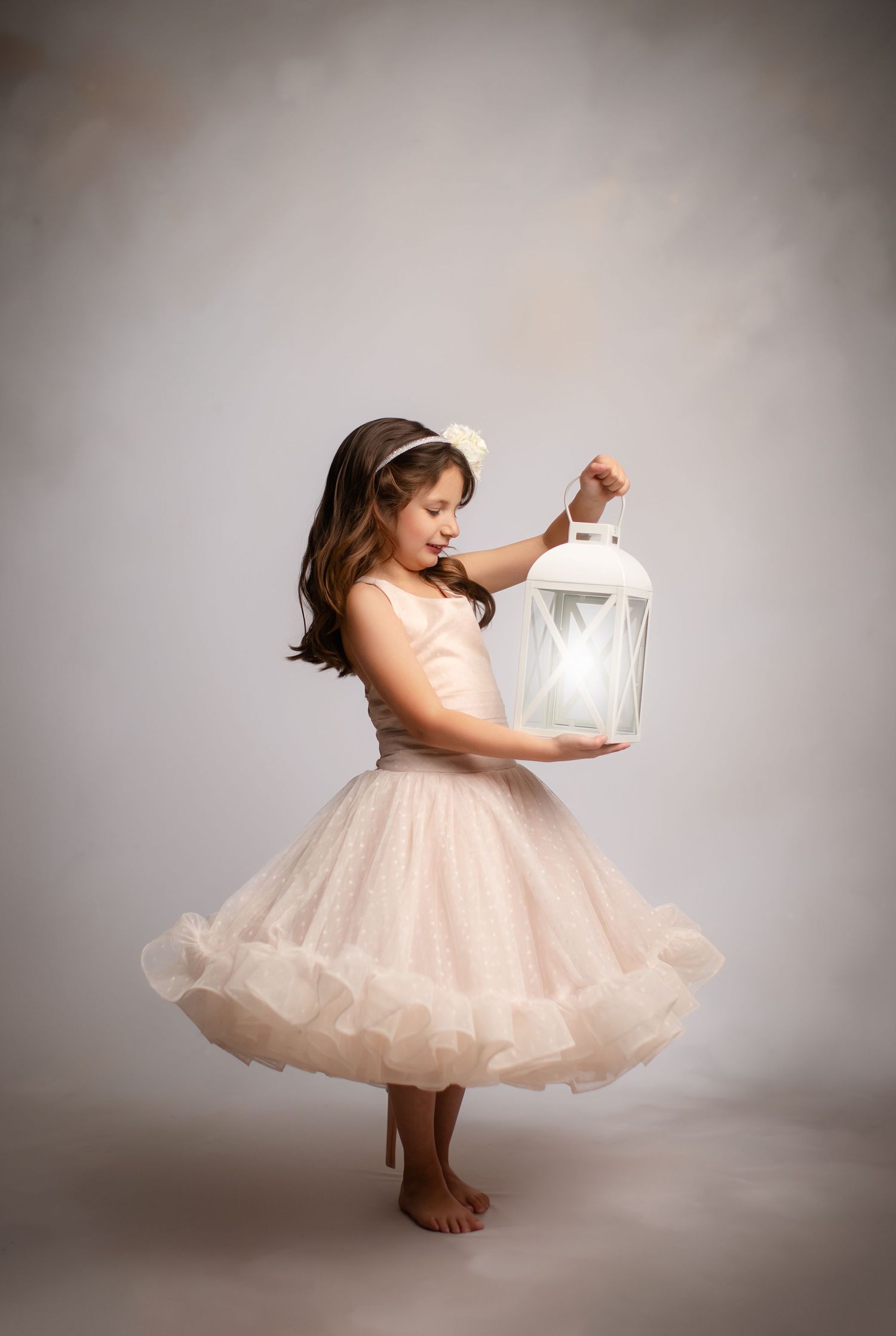 Young ballet dancer in cream tulle dress holds white lantern while posing barefoot against neutral backdrop.