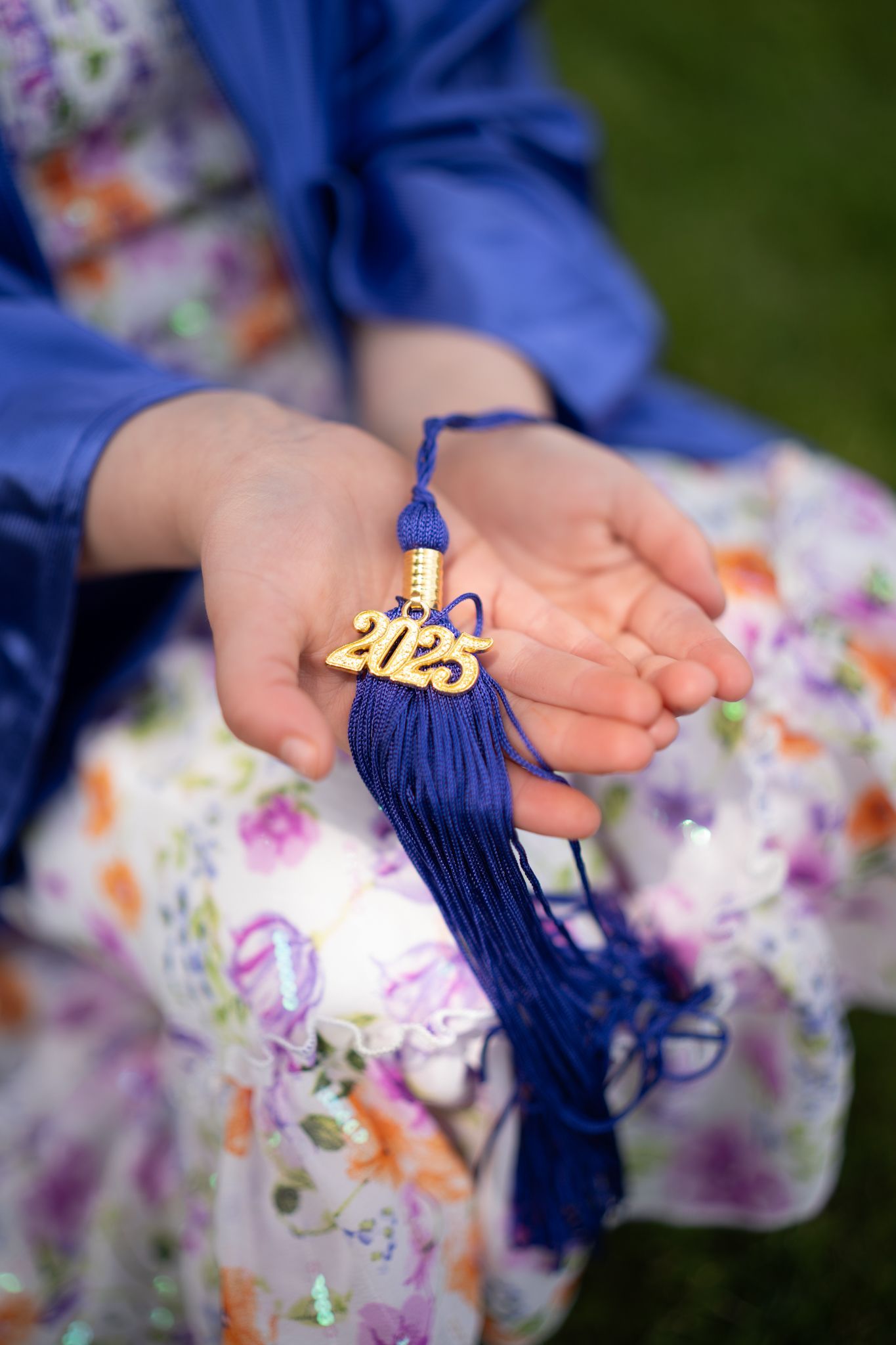 Hands holding blue graduation tassel with gold charm against floral dress fabric.
