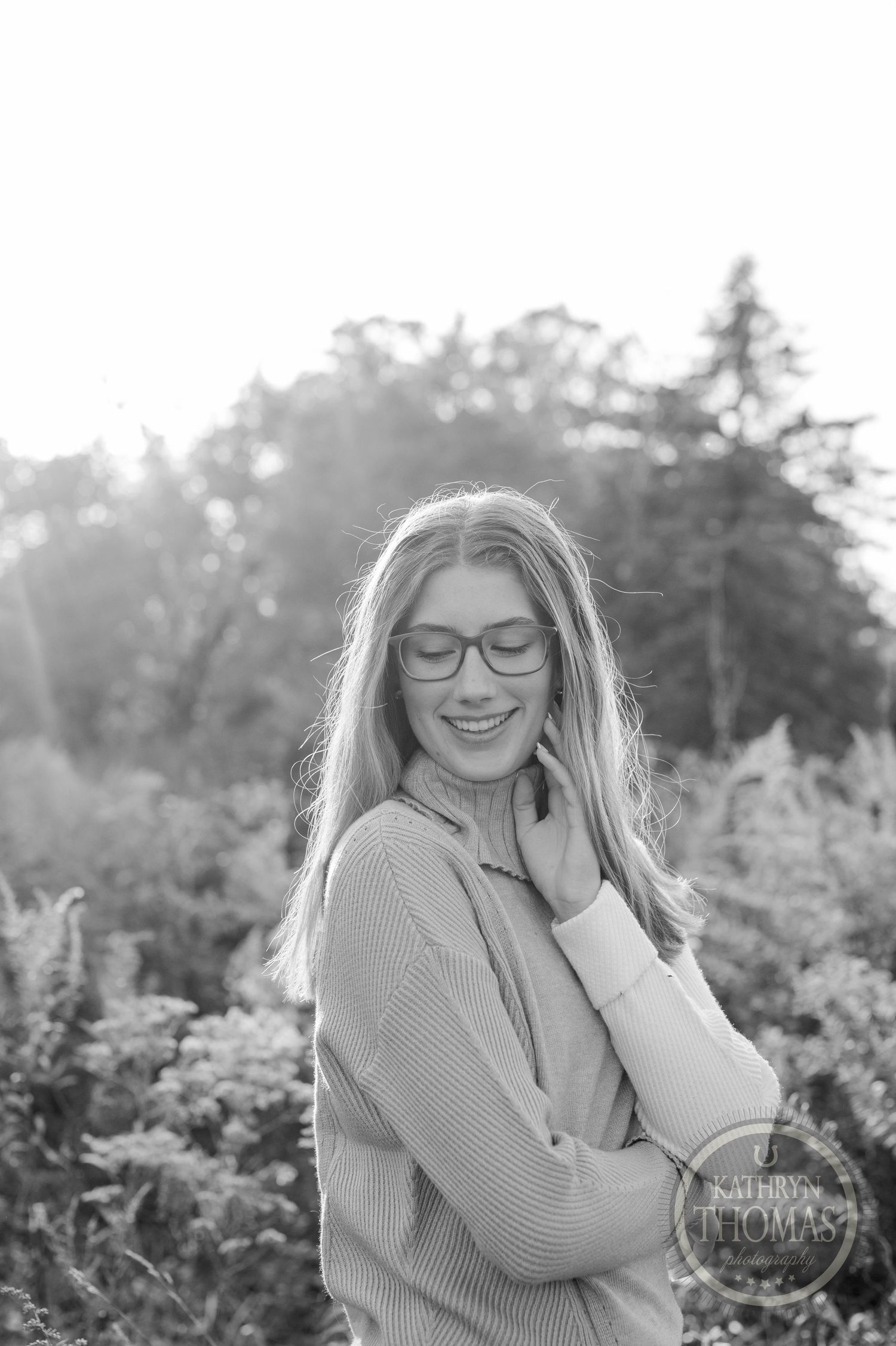 Black and white portrait in nature showing someone wearing glasses and a sweater with a gentle pose.