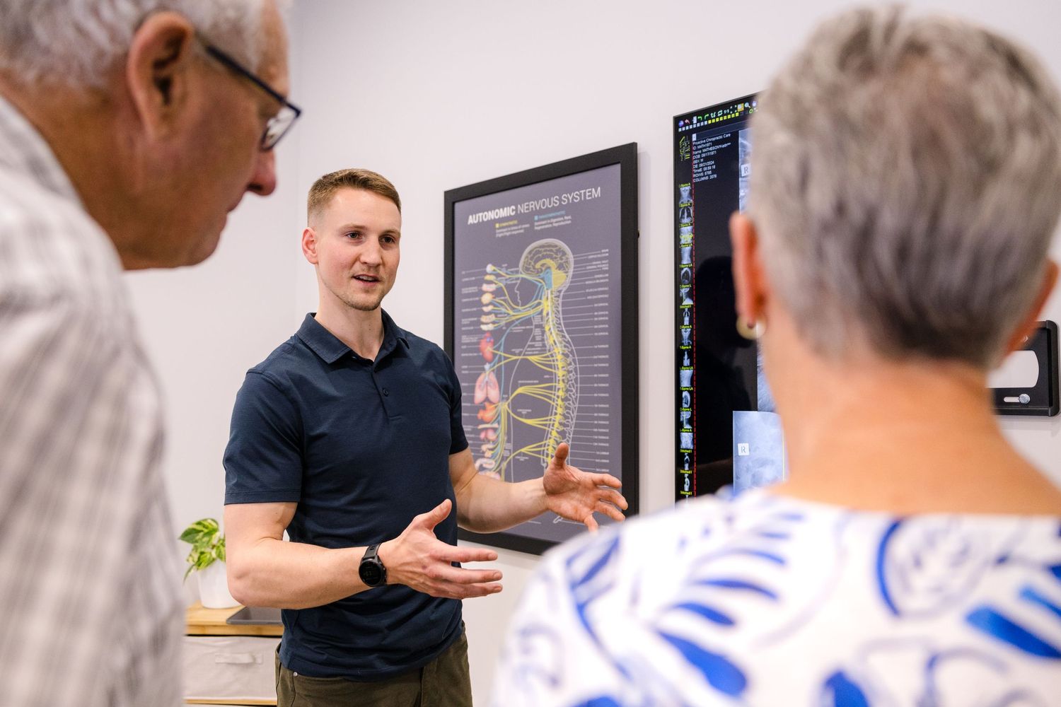 A healthcare professional points to a spinal diagram while speaking with patients in a medical office setting.