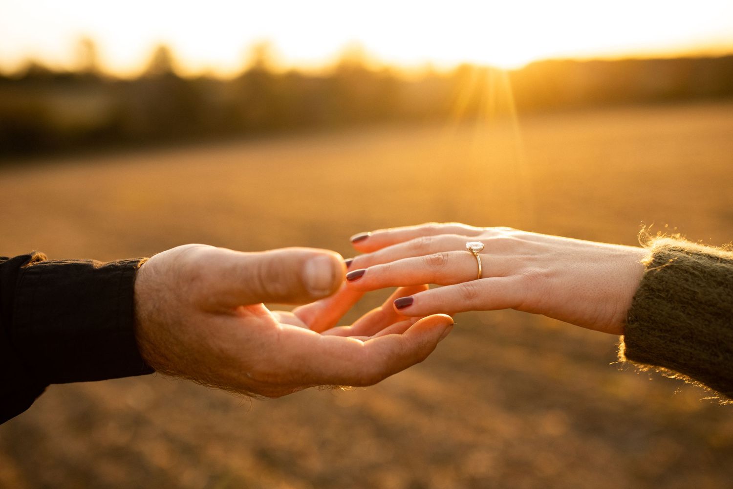 Two hands reaching towards each other against a golden sunset background, with one wearing an engagement ring.