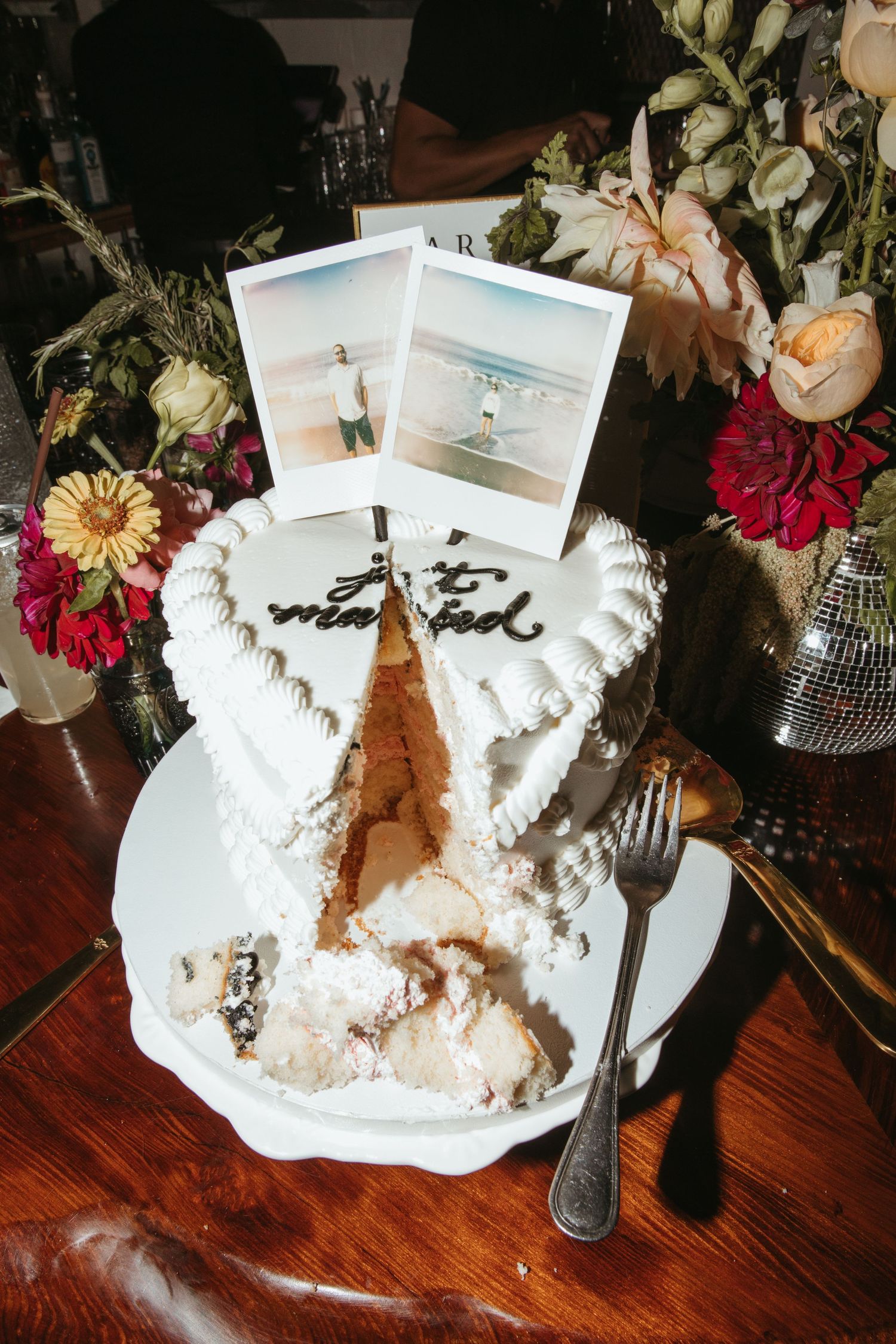 A partially eaten white frosted cake with Polaroid photos on top sits among colorful flowers on a wooden table.