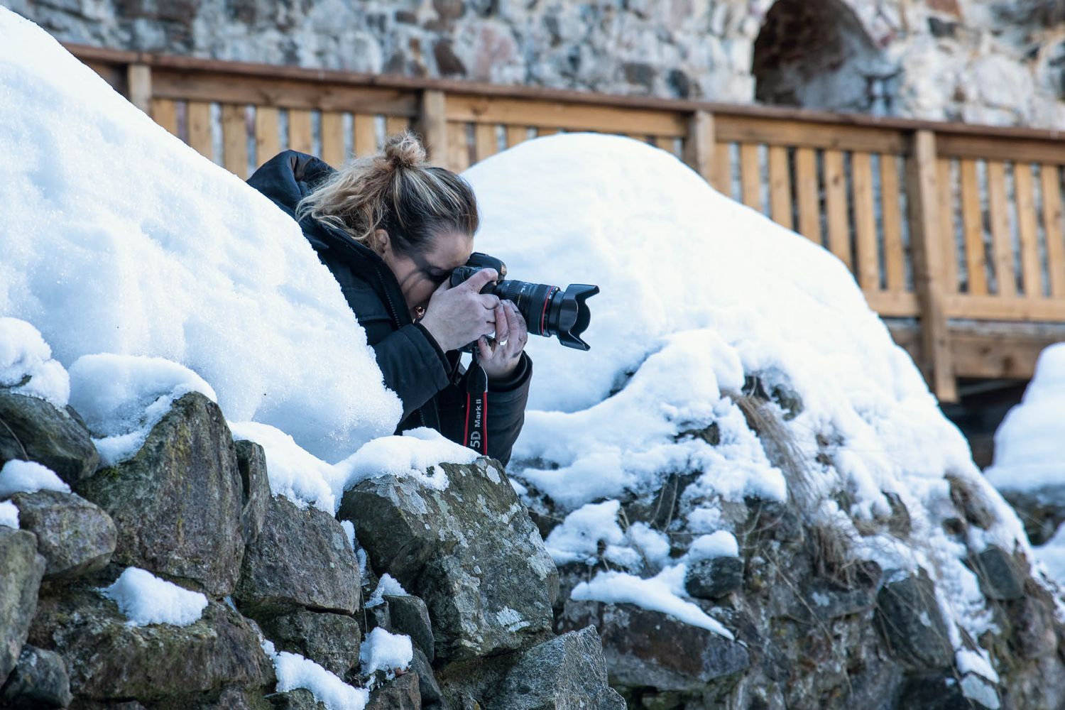 Photographer crouches behind snow-covered rocks with camera gear to capture winter scenery.