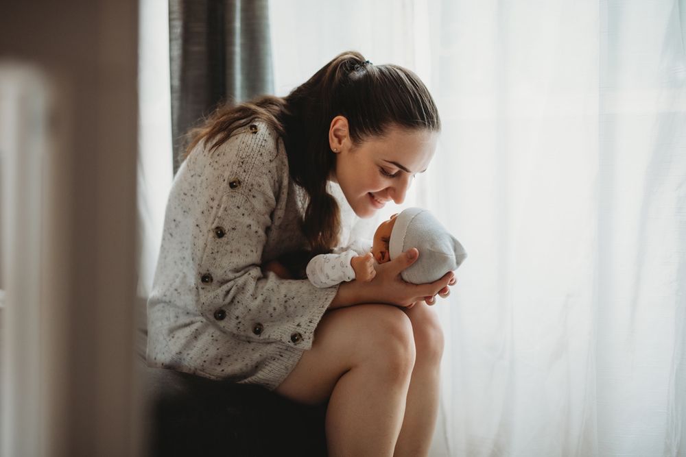 A caregiver in a knit sweater lovingly embraces a small infant by a sunlit window.