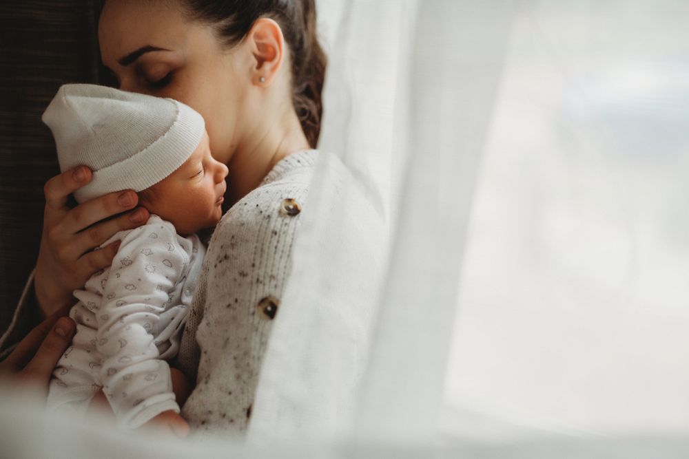 A tender moment between parent and baby by a window in soft natural light, while wearing cozy knit sweaters.