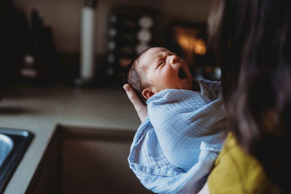 Baby in light blue outfit yawns while being held in dimly lit room.