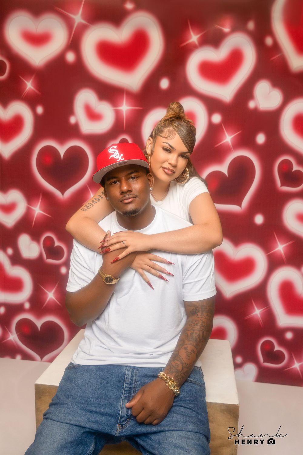 A couple in matching white shirts pose together against a backdrop of floating hearts.