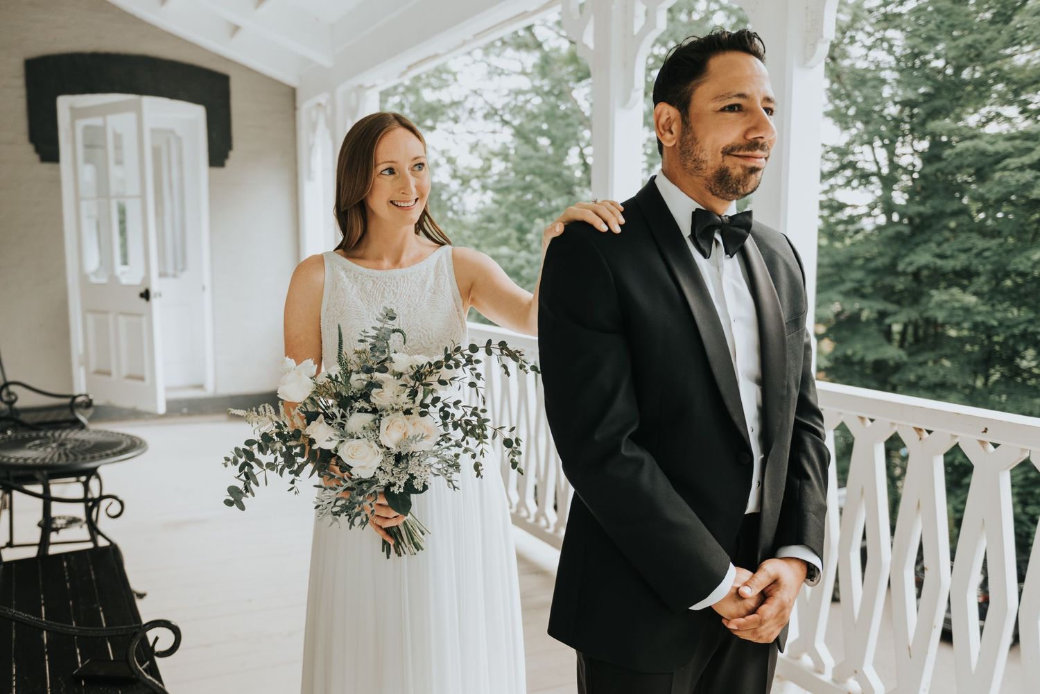 A joyful first look moment on a covered porch as a couple shares a laugh during their wedding day celebration.