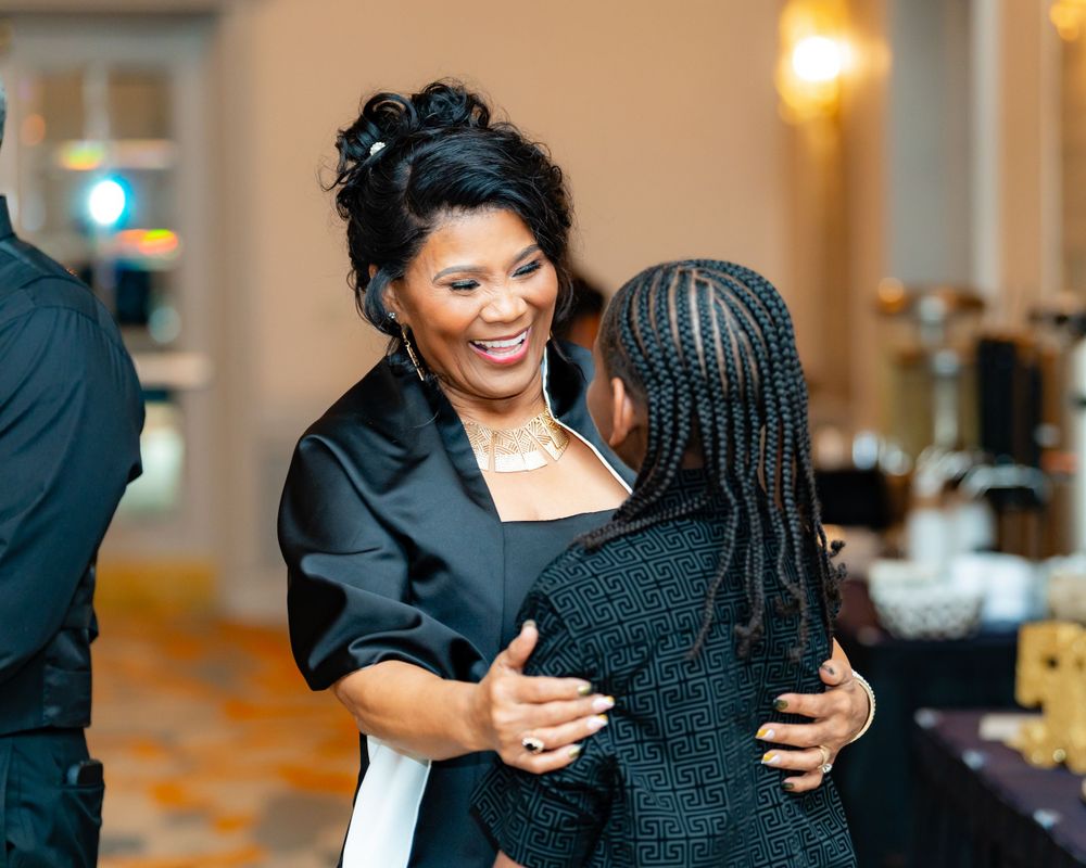 Two people share a warm embrace while laughing together at a formal indoor gathering.