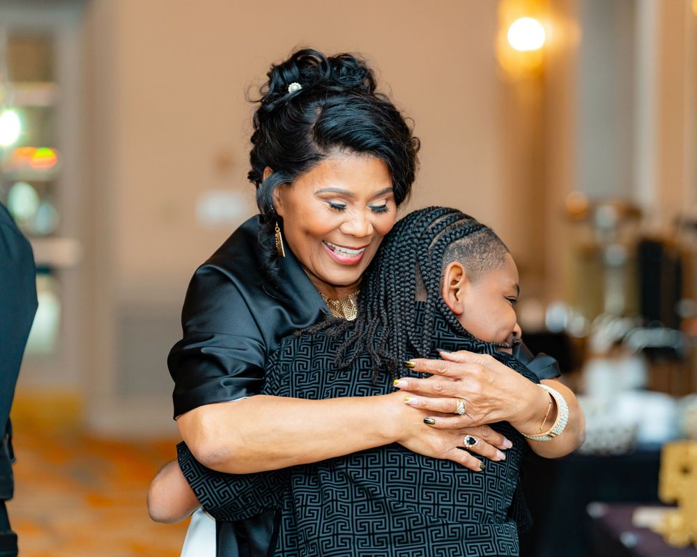 Two people share a joyful embracing moment at an indoor event wearing formal attire.