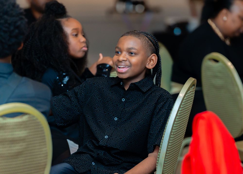 Young person in a black button-up shirt smiles while seated on a green chair at an event.
