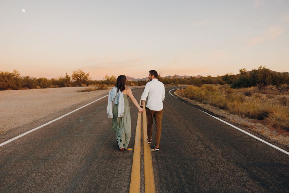 A couple holding hands while walking down a long desert highway.
