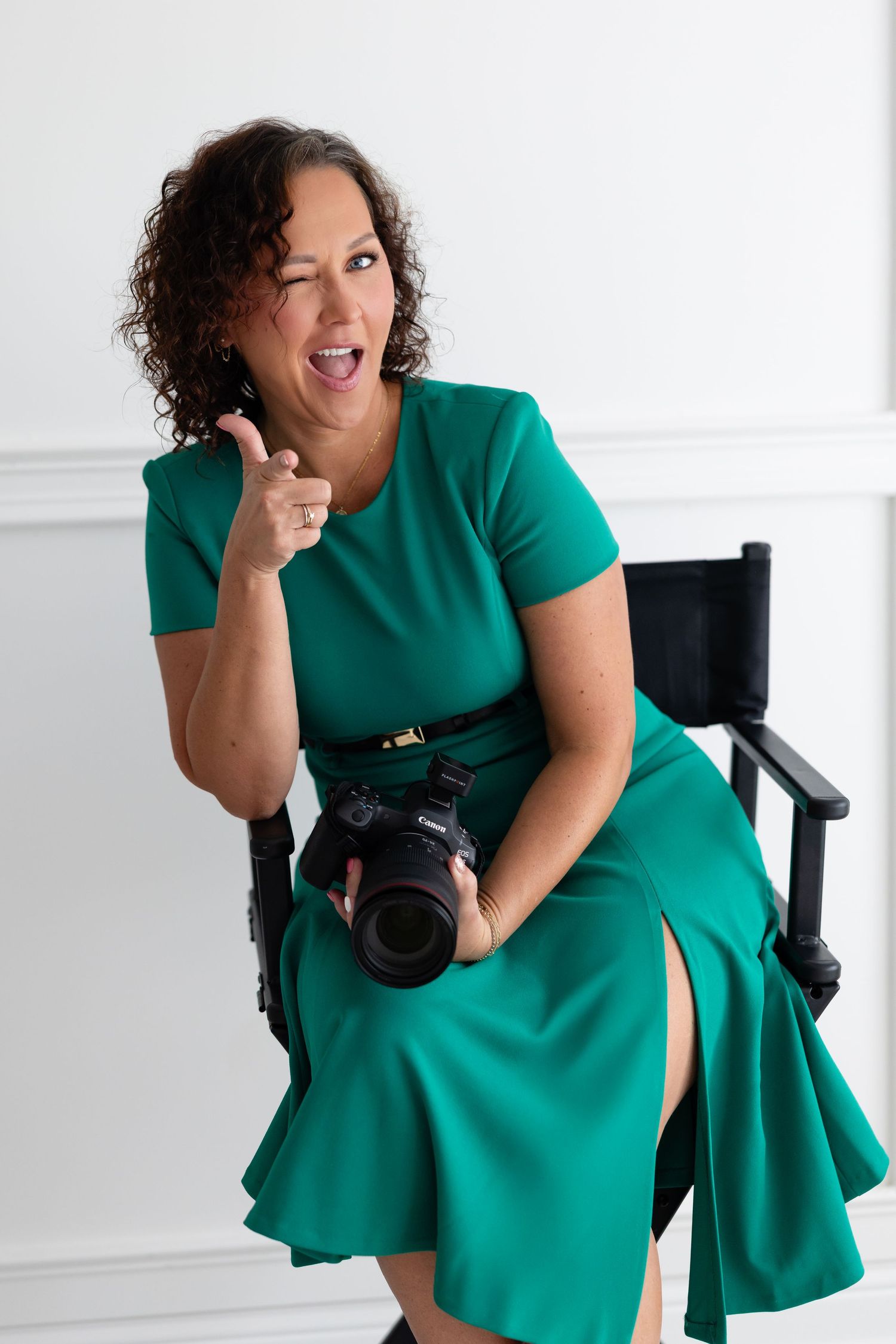 Professional photographer in emerald green dress sits in director's chair holding DSLR camera while making winking gesture.