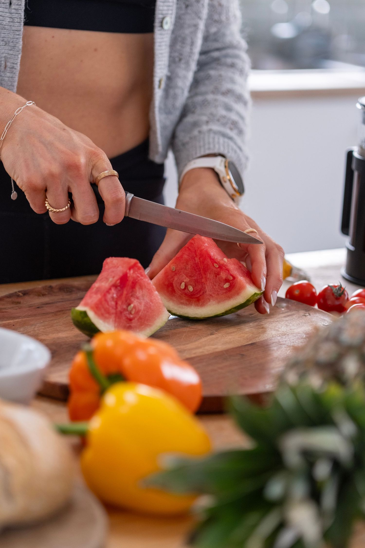 Hands cutting fresh watermelon on wooden board with colorful produce in background.
