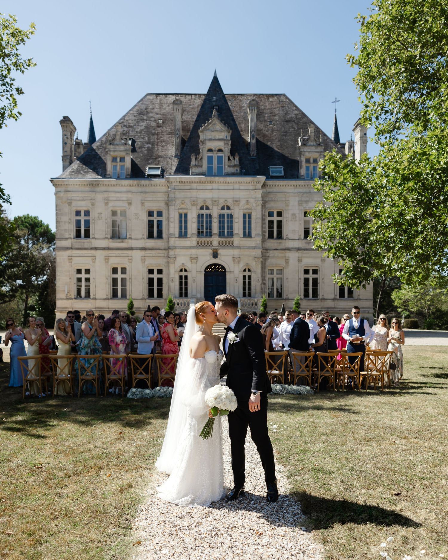 A couple celebrates their wedding in front of a grand French chateau on a sunny summer day with guests looking on.