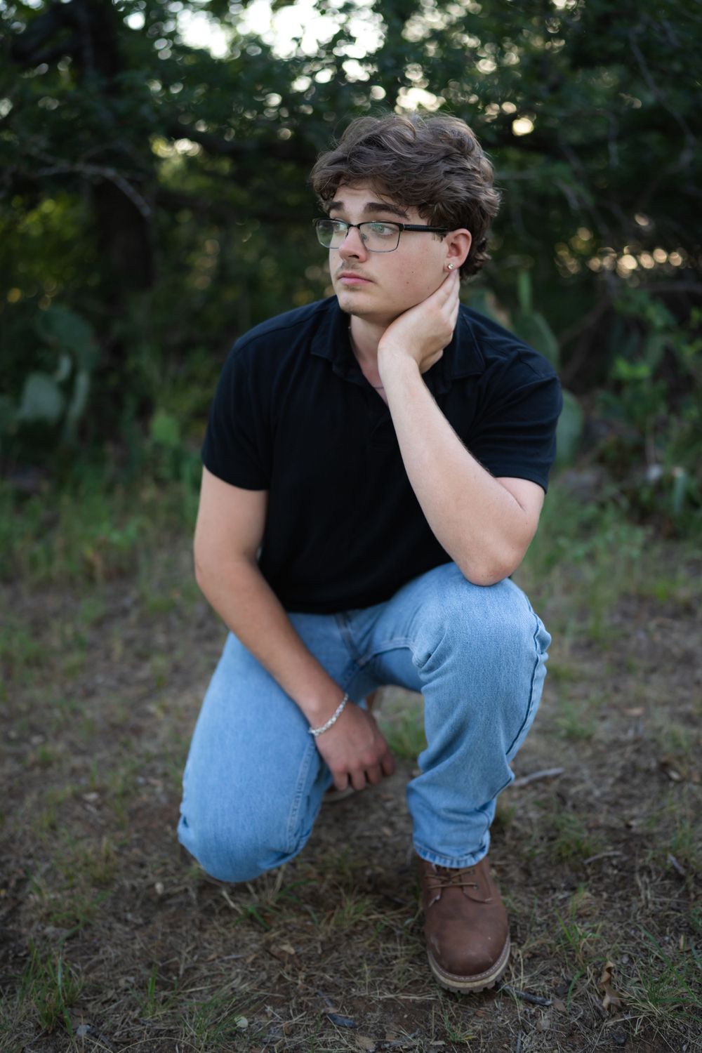Someone in glasses wearing a black polo shirt and jeans poses crouched in an outdoor setting at dusk.