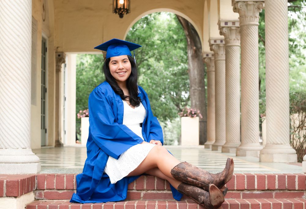 Graduate in blue cap and gown sits on stone steps with brown boots against classical archway backdrop in San Antonio, Texas