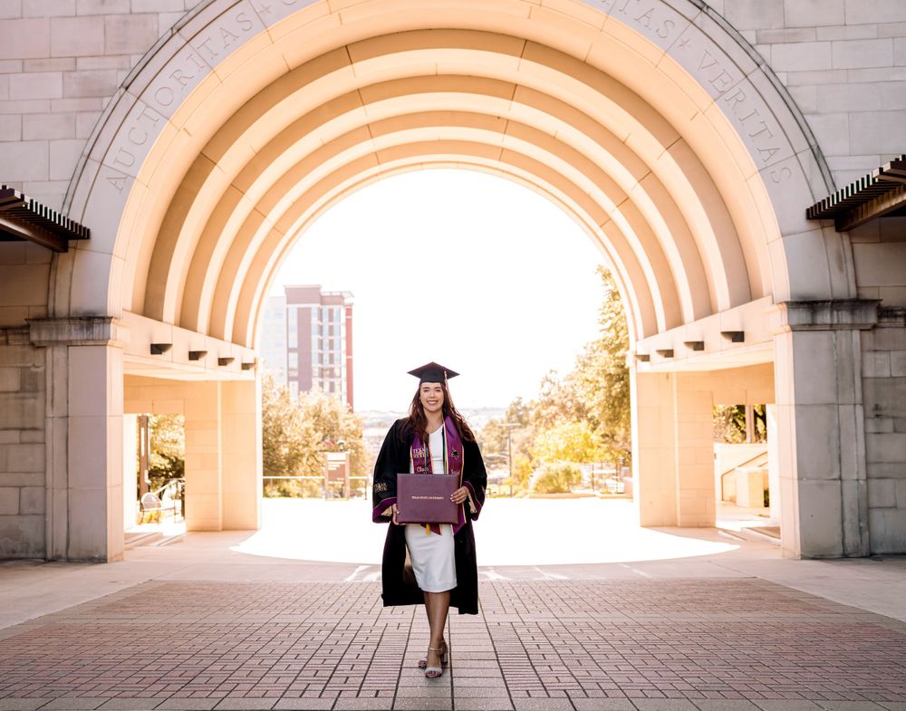 Graduate in cap and gown poses beneath an arched passageway at a university campus during graduation.
