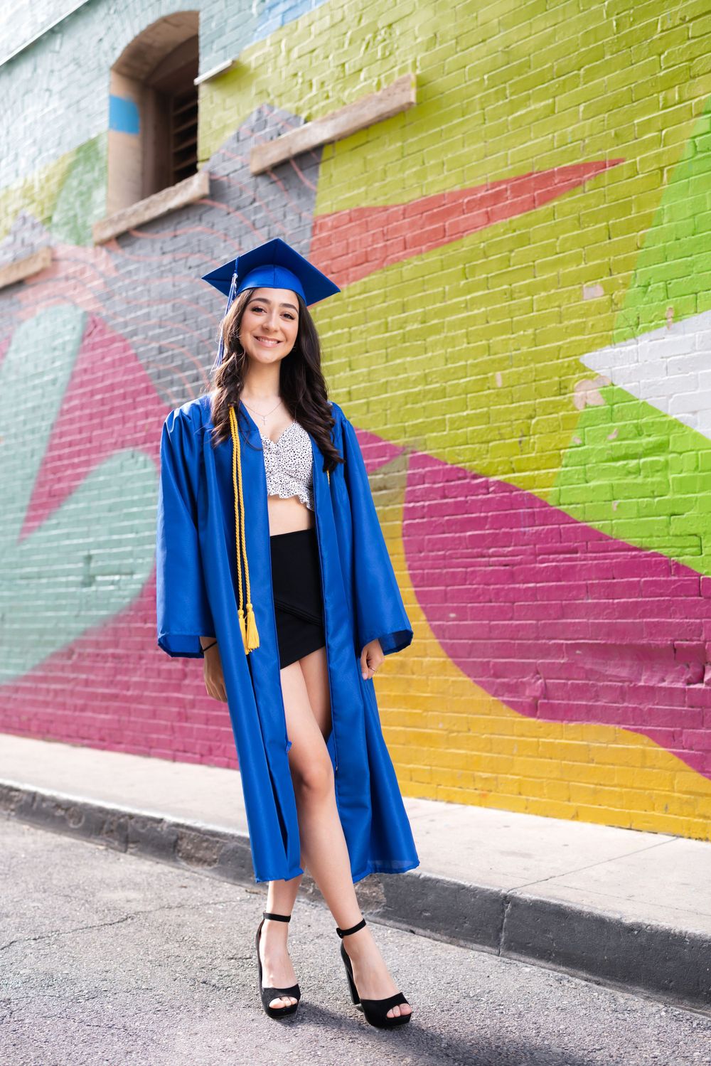 Graduate in a blue cap and gown stands in front of a colorful geometric mural painted on brick wall.