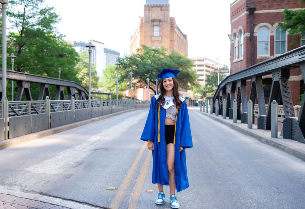 Graduate in bright blue graduation gown and cap standing on a bridge with urban architecture in the background.
