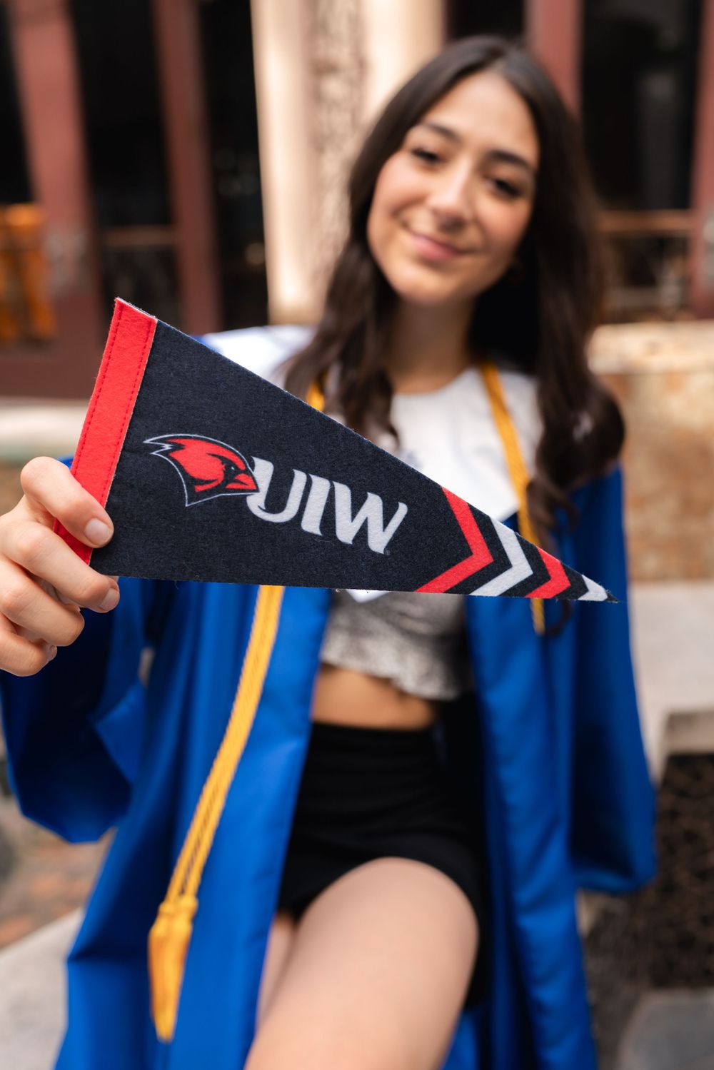 Someone in graduation attire holds a UIW university pennant while wearing a blue commencement gown and white top.