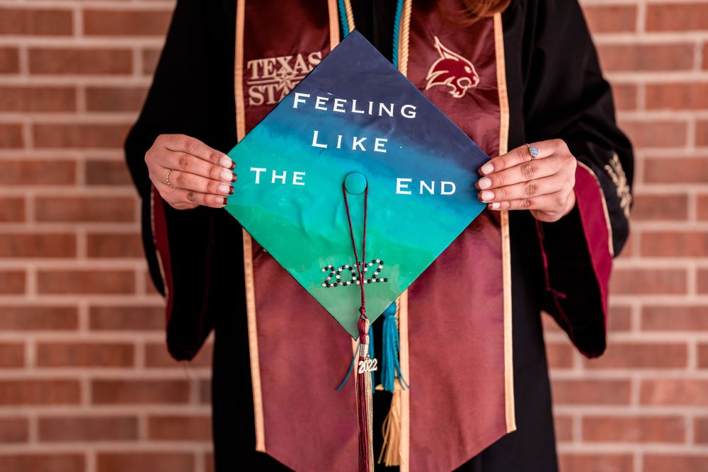 A graduation cap decorated with turquoise ombre design and text reading 'Feeling like the end' against a brick wall.