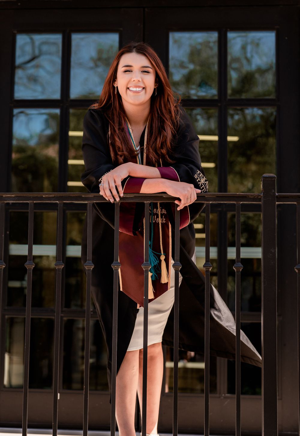 Someone in a black cardigan leans against a metal railing outdoors with a bright smile on a sunny day.